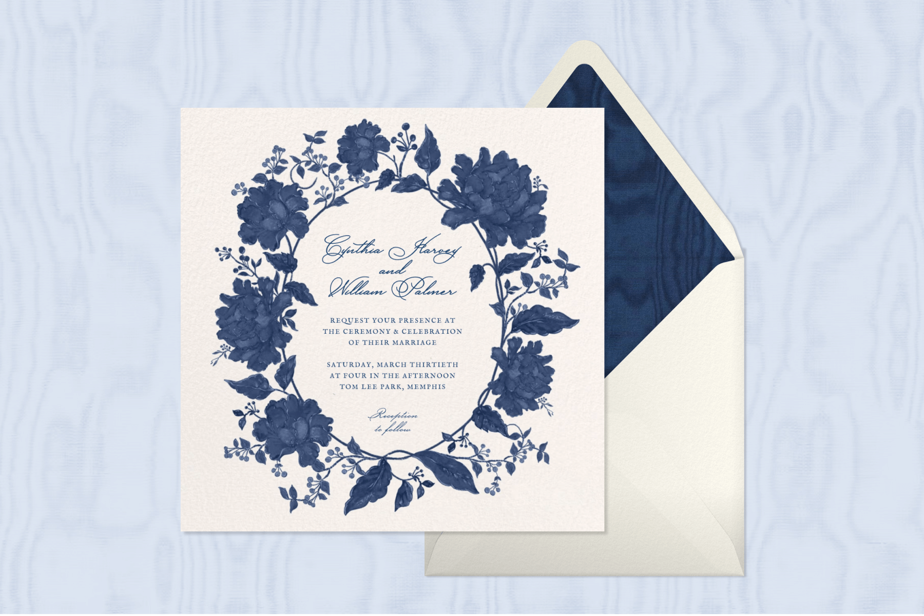 A white wedding invitation with navy script text and a navy illustrated floral wreath border with a white envelope and navy fabric envelope liner.