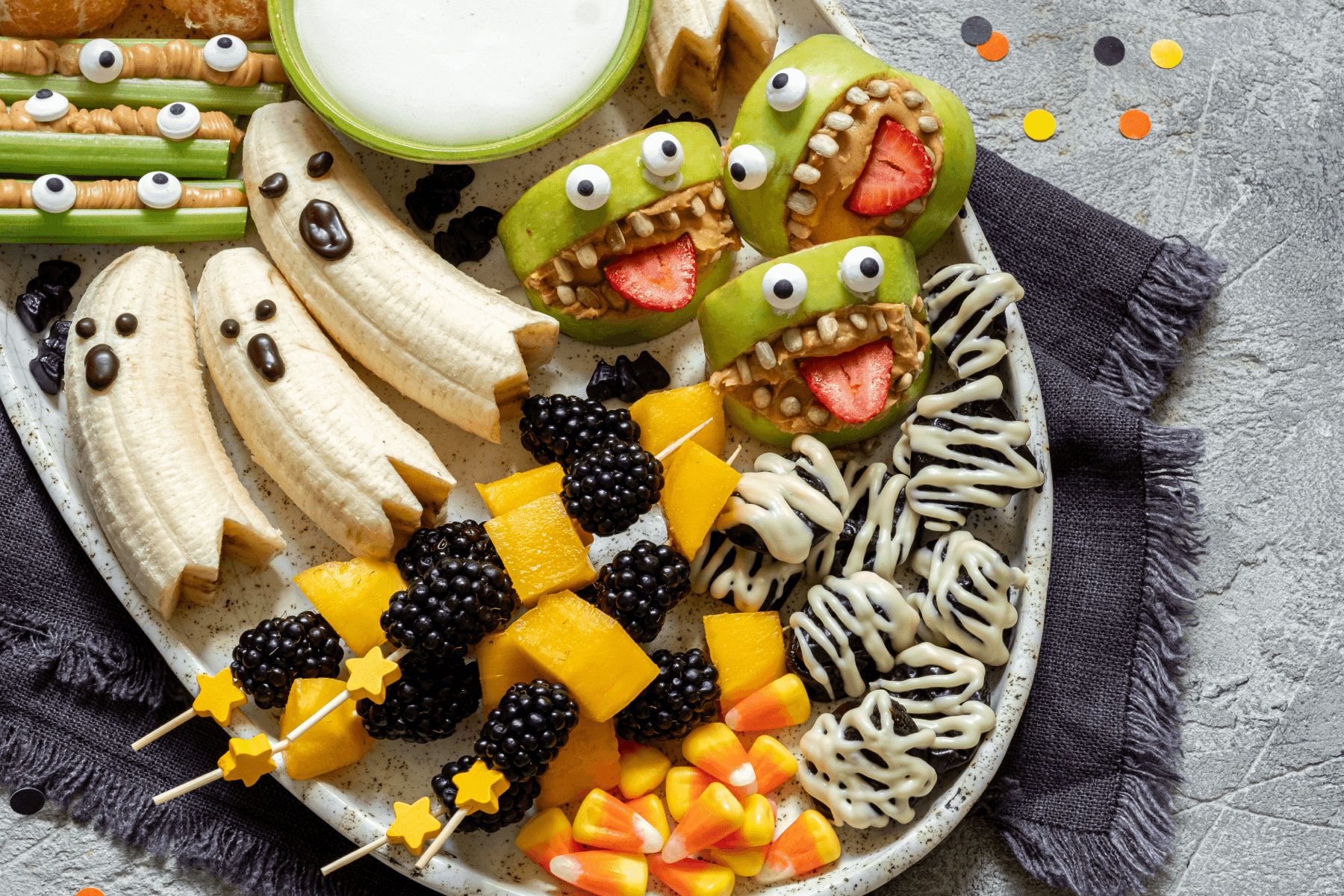 A Halloween snack platter with banana ghosts, green apple monsters with strawberry tongues and candy eyes, celery sticks with peanut butter, blackberry and mango skewers, candy corn, and white chocolate mummy treats on a dark napkin over a grey surface scattered with confetti.
