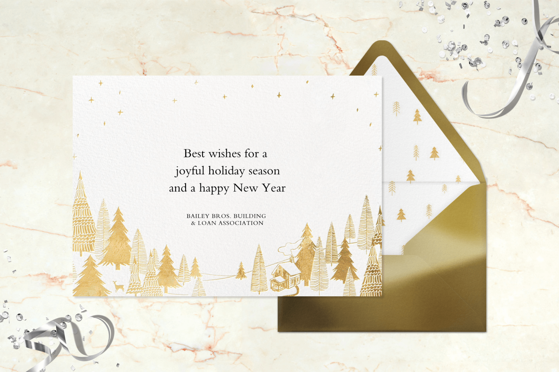 A white holiday card has gold evergreen trees, a cabin, and deer along the bottom with a gold envelope.