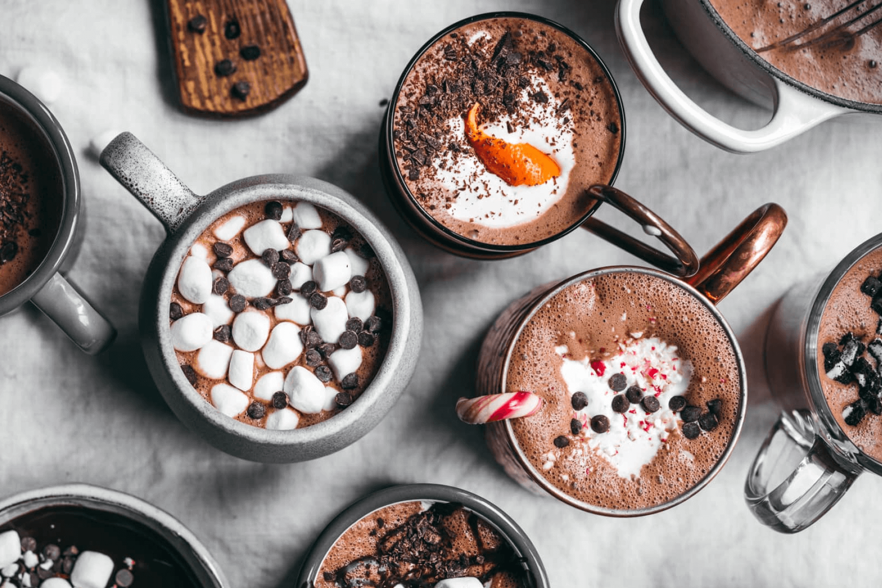Several mugs of hot chocolate with various sweet toppings like marshmallows, chocolate chips, and candy canes.