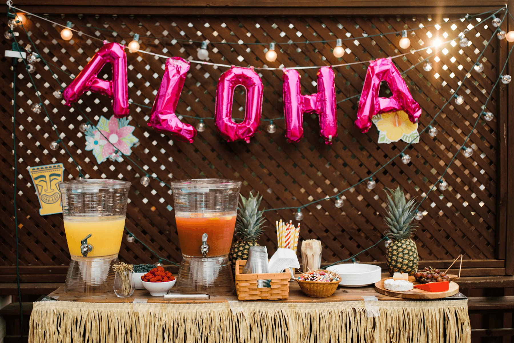 An outdoor table with grass trim has drink dispensers and pineapples and a pink balloon ‘ALOHA’ banner