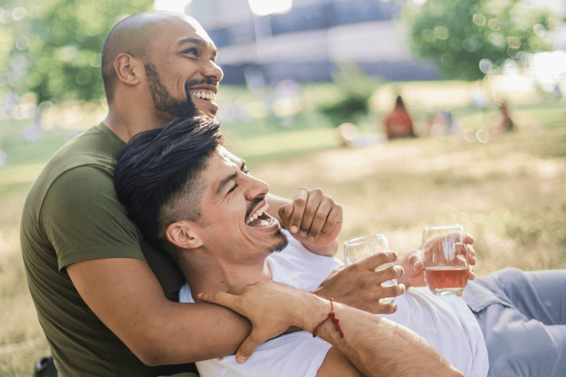 A man lies down on his partner’s lap as they laugh together, holding drinks and enjoying themselves in a park. 