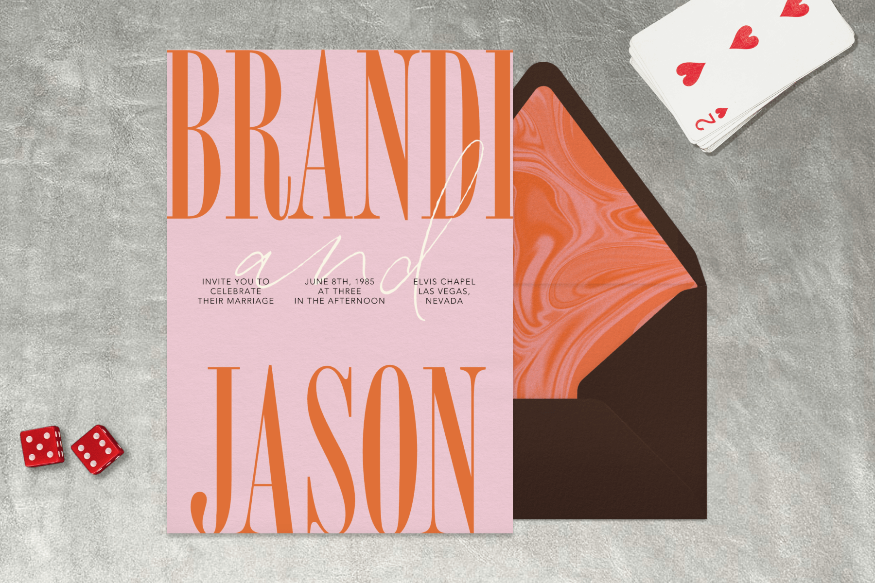 A wedding invitation with a pink background and orange text paired with a brown envelope and orange swirl envelope liner on a silver background with dice and playing card accents.