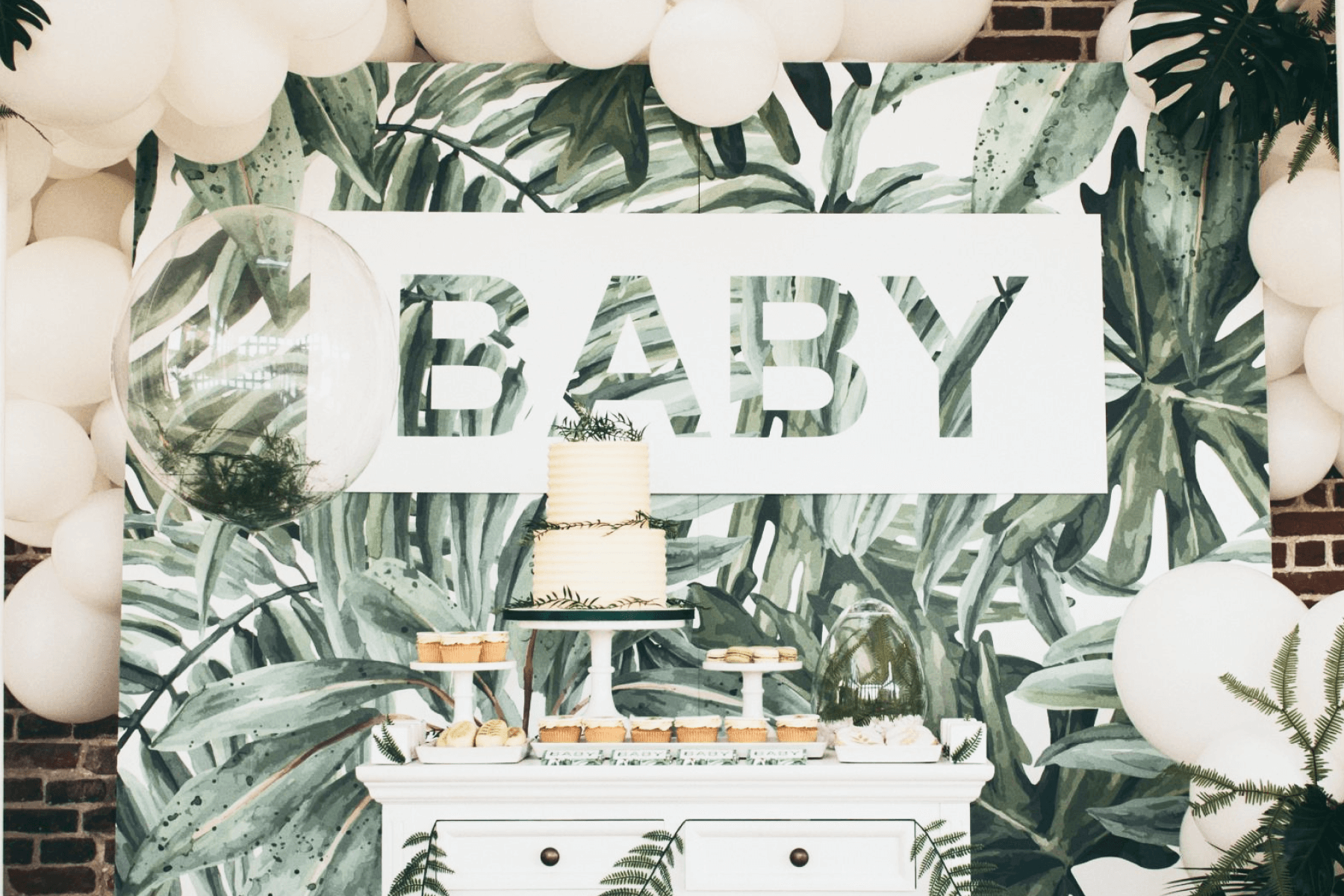 A baby shower dessert display with a two-tiered cake and a backdrop printed with tropical plant fronds and the word “BABY” spelled out in the center.