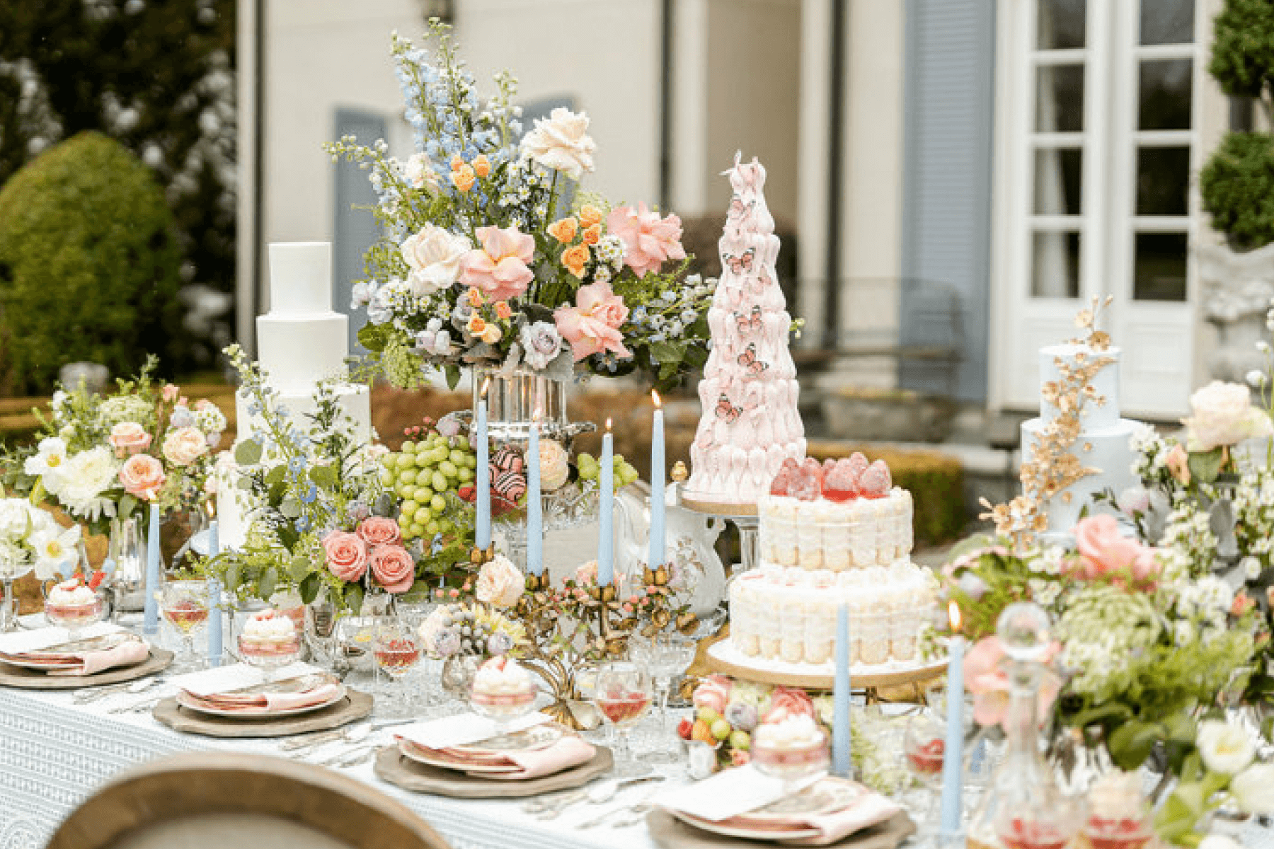 An outdoor banquet table is decorated for what appears to be a summer-y wedding or celebratory event with plenty of pastel floral arrangements (roses), grapes, tiered cakes with fruit and butterflies, and light blue taper candles.