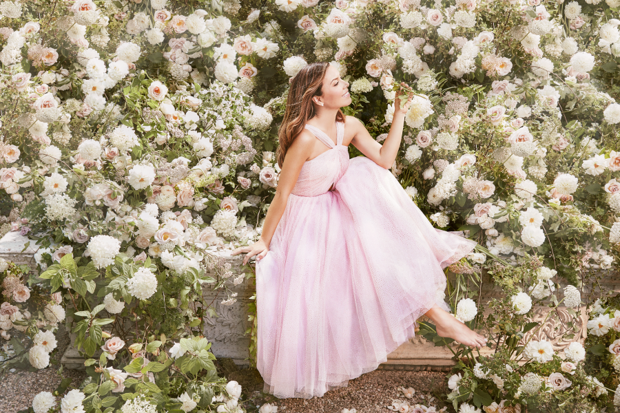 Fashion designer Monique Lhuillier wears a light pink gown and sits on a bench framed by bushes of white roses.