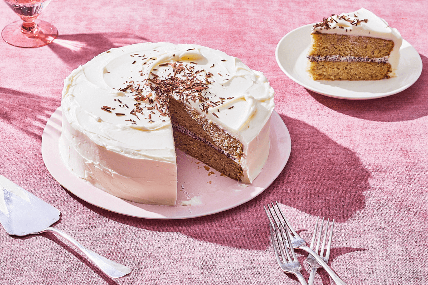 A carrot cake with white frosting and a slice cut out sits next to the missing slice on a pink tablecloth beside silverware.