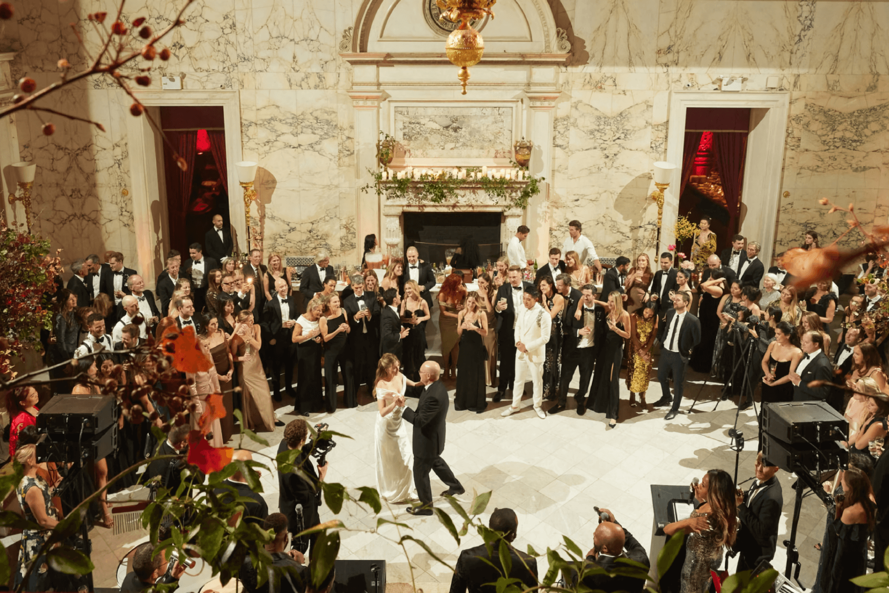 A father and daughter share a wedding dance surrounded by guests in an opulent room with marble walls and tall ceilings.