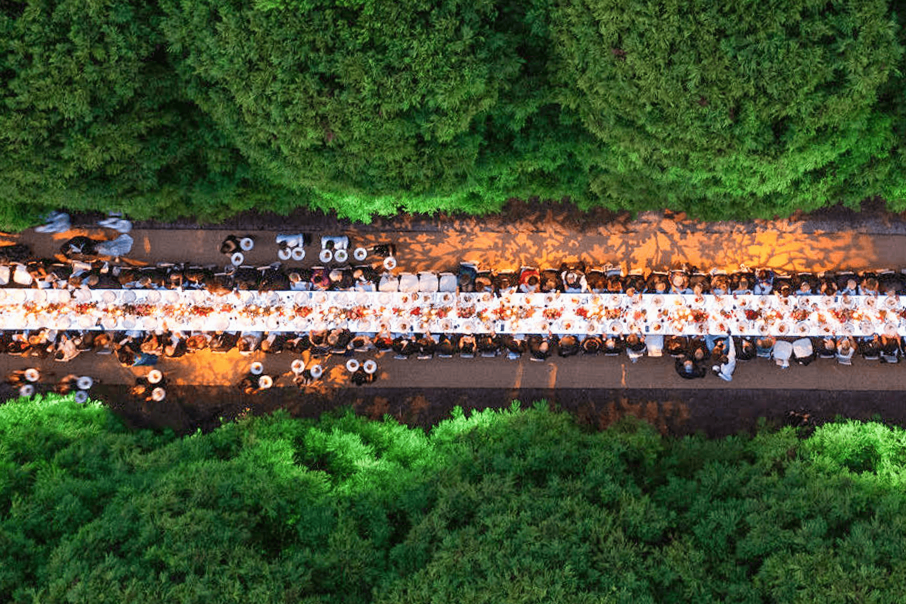 Viewed from above, a very long white wedding banquet table with servers and guests is set up on a road surrounded by green trees.