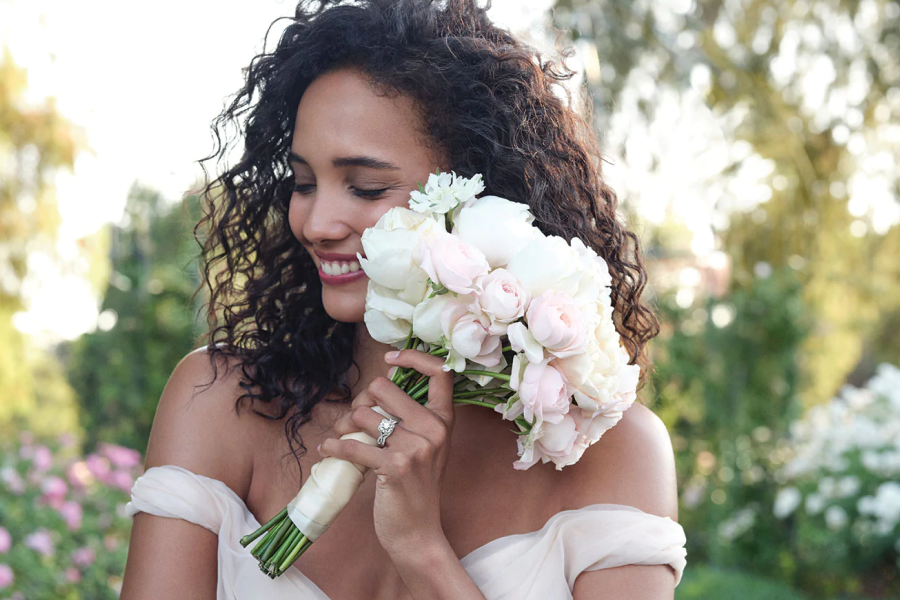 Portrait of a bride holding a bouquet of pink and white peonies, with an off-white silk ribbon wrapped around the stems, close to her face.