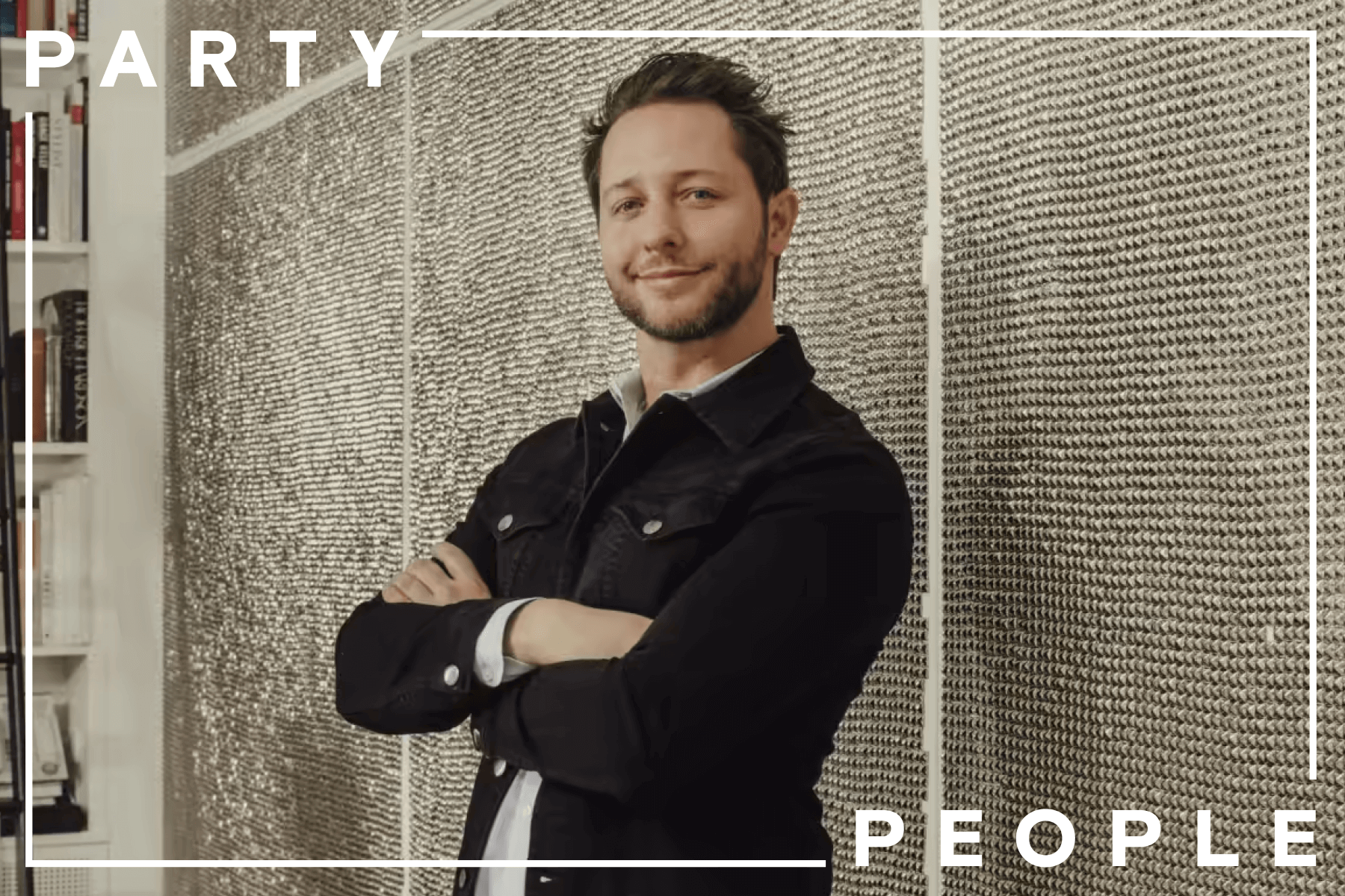 Derek Blasberg, wearing a dark denim jacket, stands in front of a silver wall smiling with his arms folded.