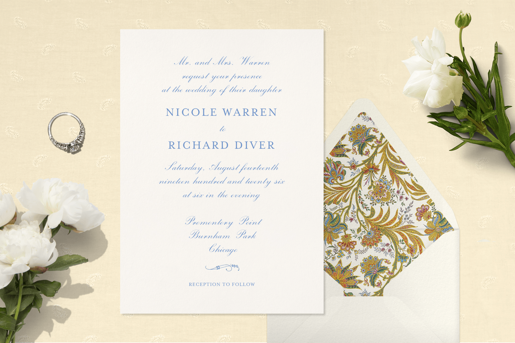 On a tan backdrop, a white wedding invitation with blue lettering sits next to a white envelope with floral patterned lining. The backdrop also features white flowers and an engagement ring.