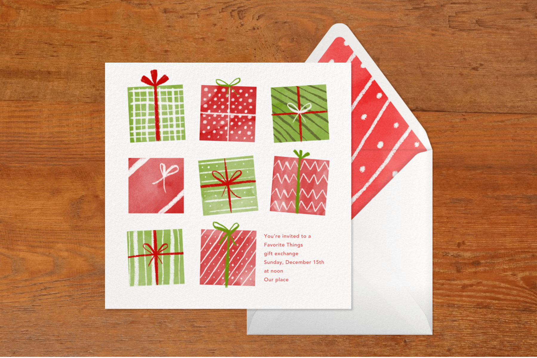 A square white invitation featuring red and green patterned gift boxes, on top of a white envelope with a red and white striped and dotted liner, set against a dark wood backdrop.