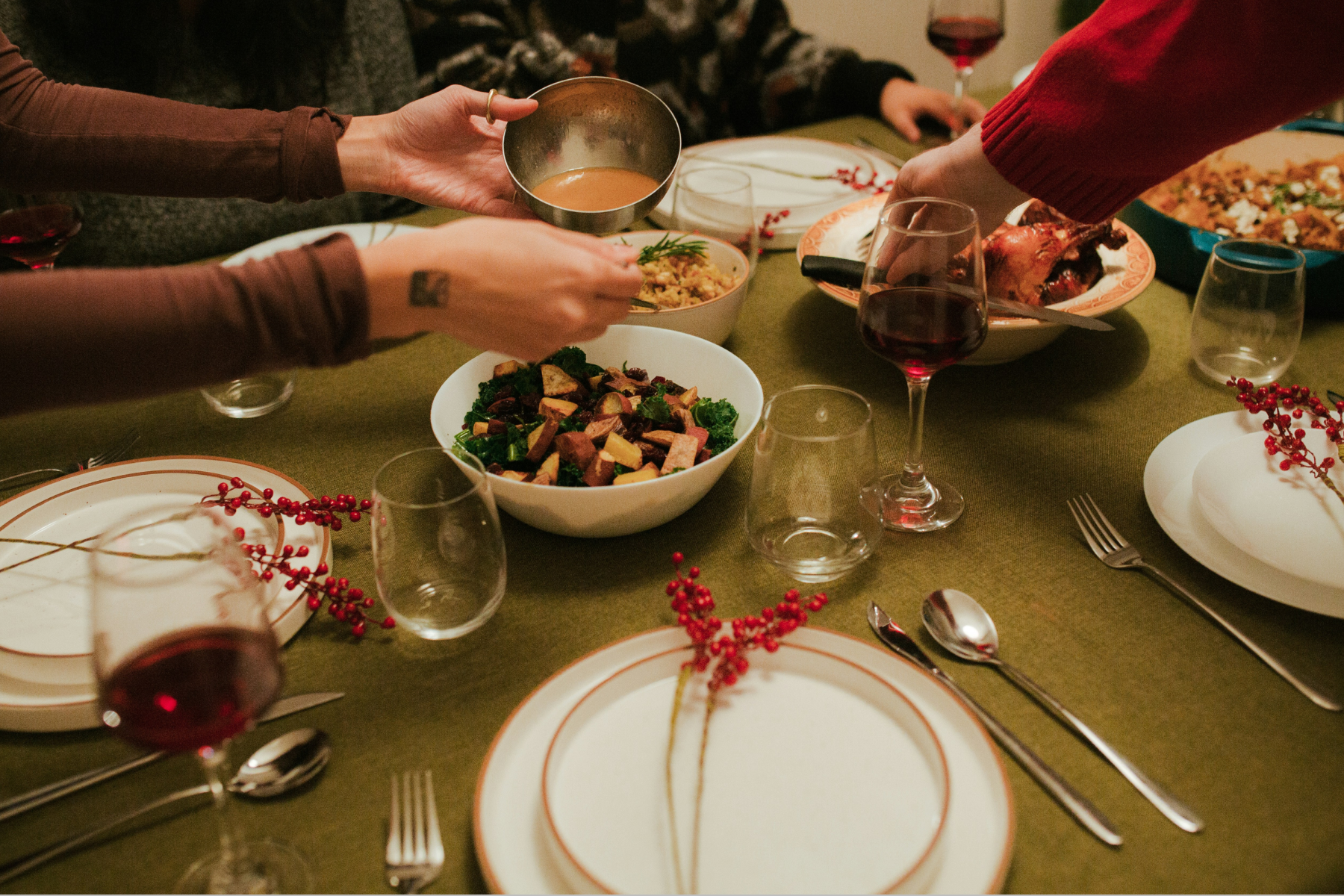 A Thanksgiving table with greens, gravy, and stuffing on a green tablecloth, surrounded by white plates, wine glasses, and sprigs of red berries. 