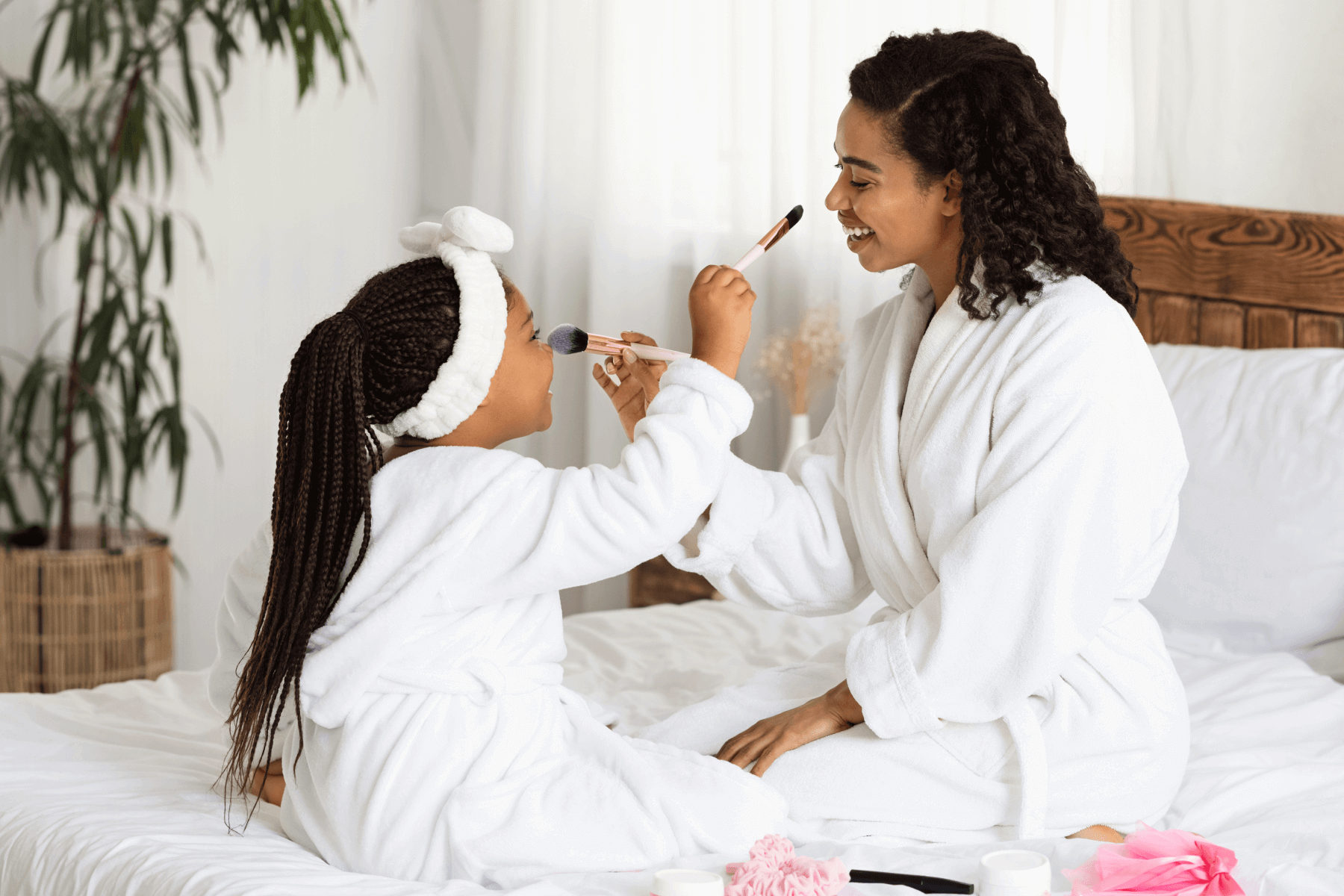 A mother and daughter in white robes on a bed play with makeup brushes.