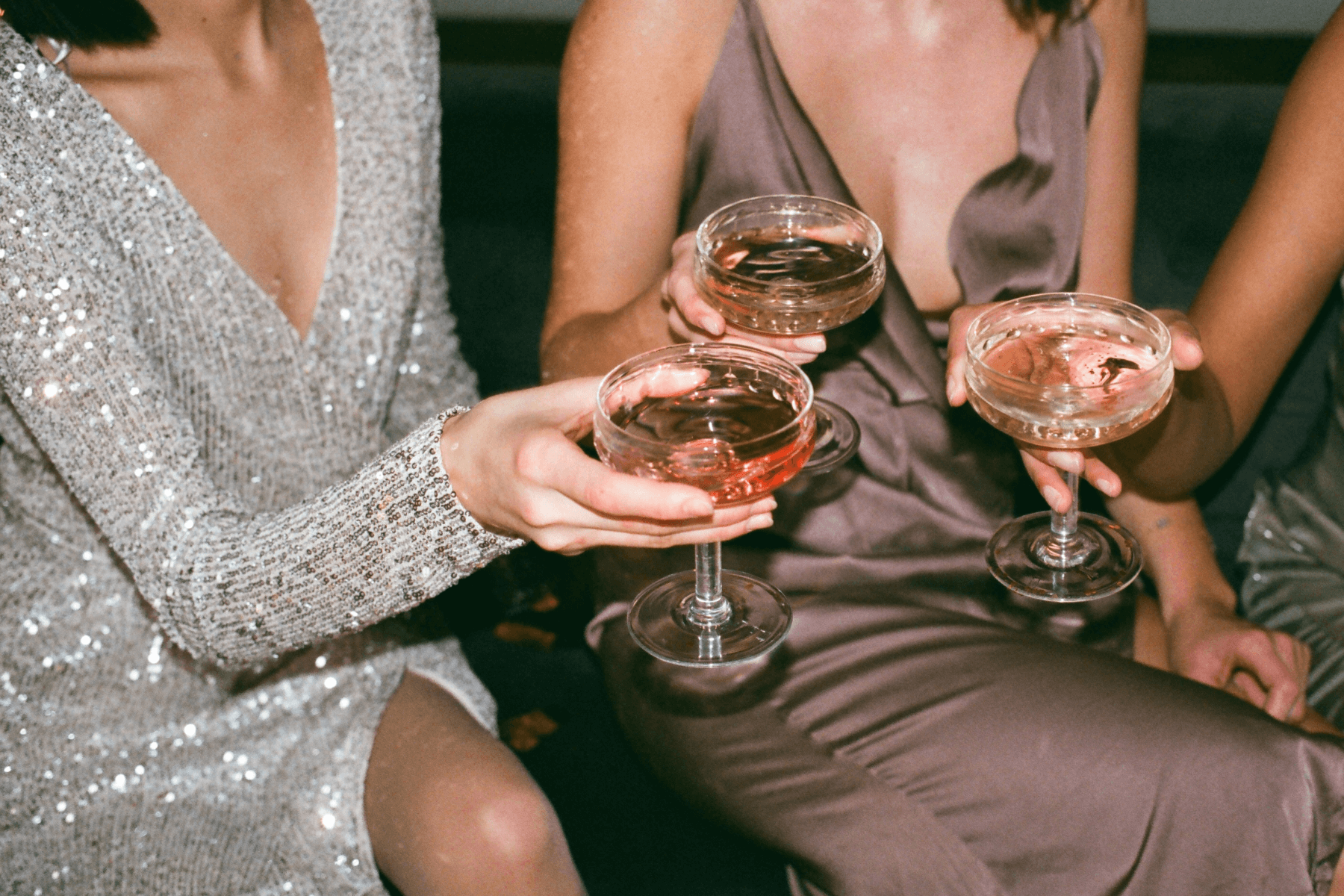 Three women with faces out of frame in formal attire clink coupe glasses.