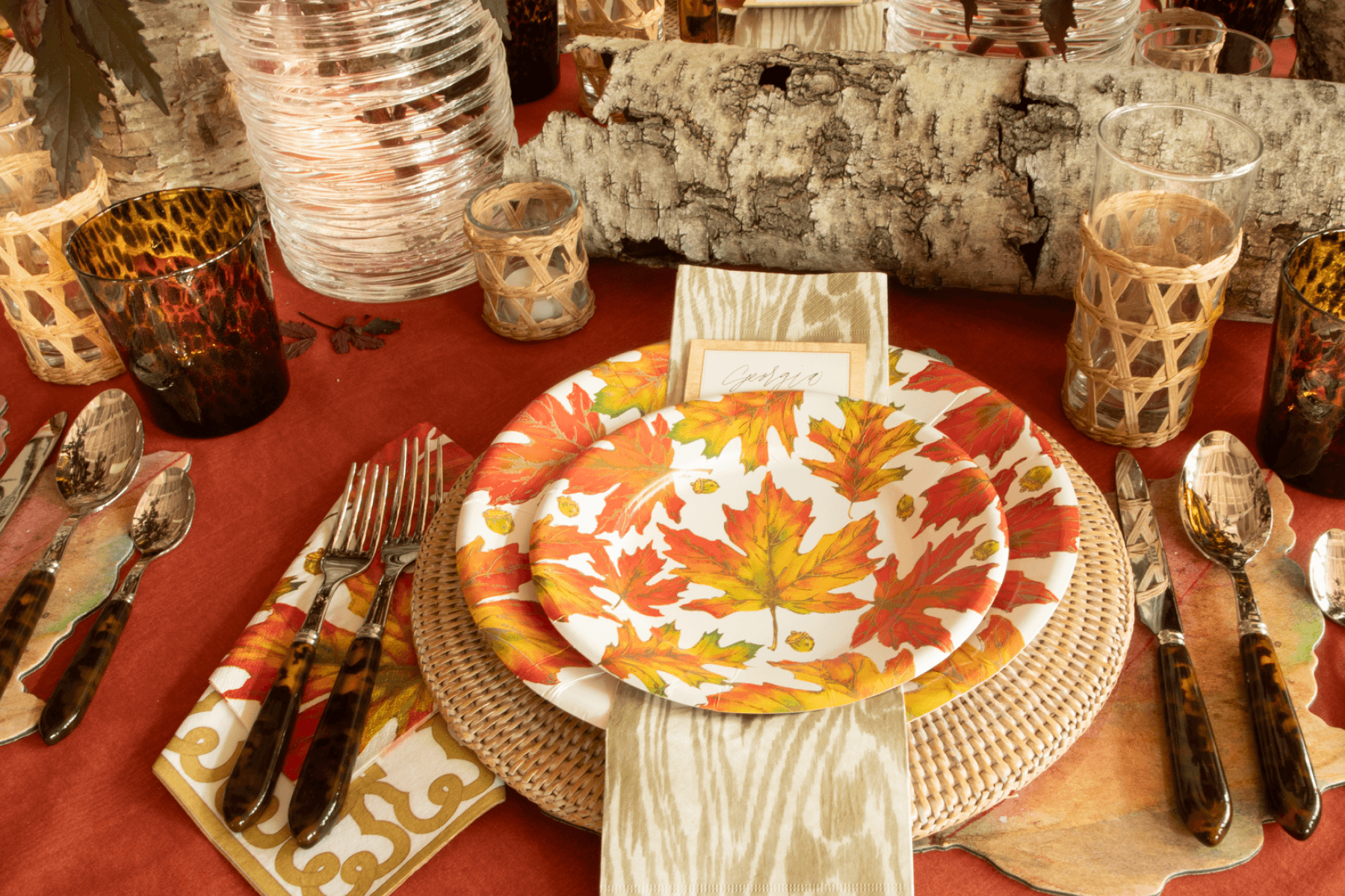 A table set with autumn leaf paper plates, tortoiseshell cutlery, and decorative birch logs.