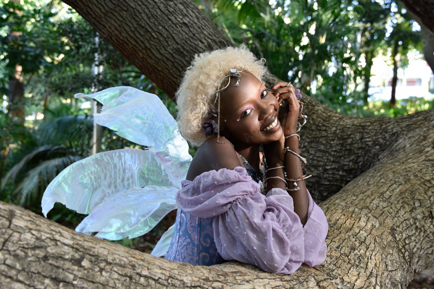 A person in a fairy costume with iridescent wings, a lilac puff-sleeve top, a jeweled headpiece, and stacked silver bracelets smiles while leaning on a large tree branch in a leafy garden.