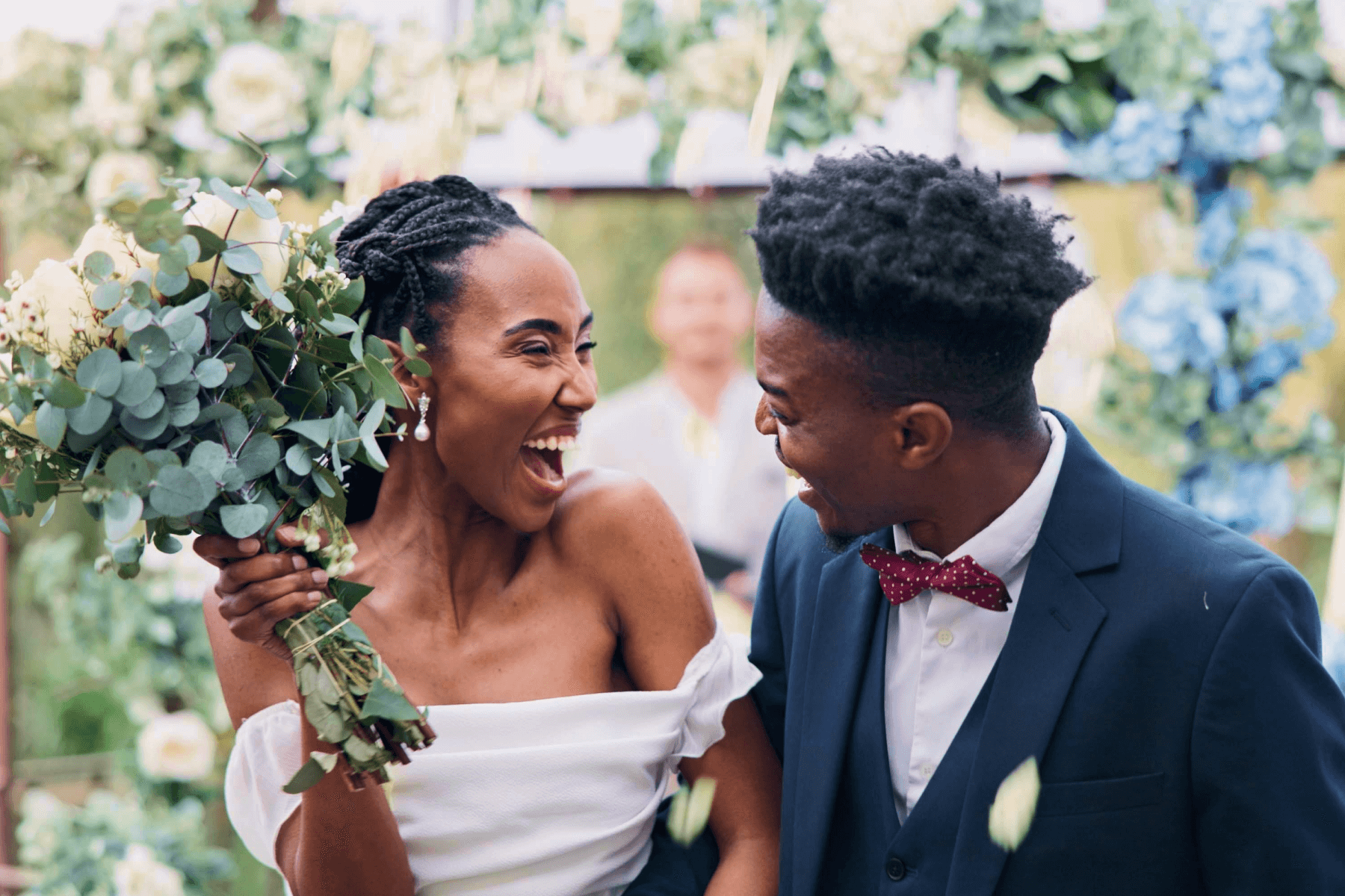 A bride and groom looking at each other and smiling while the bride holds up a bouquet of flowers and greenery. 