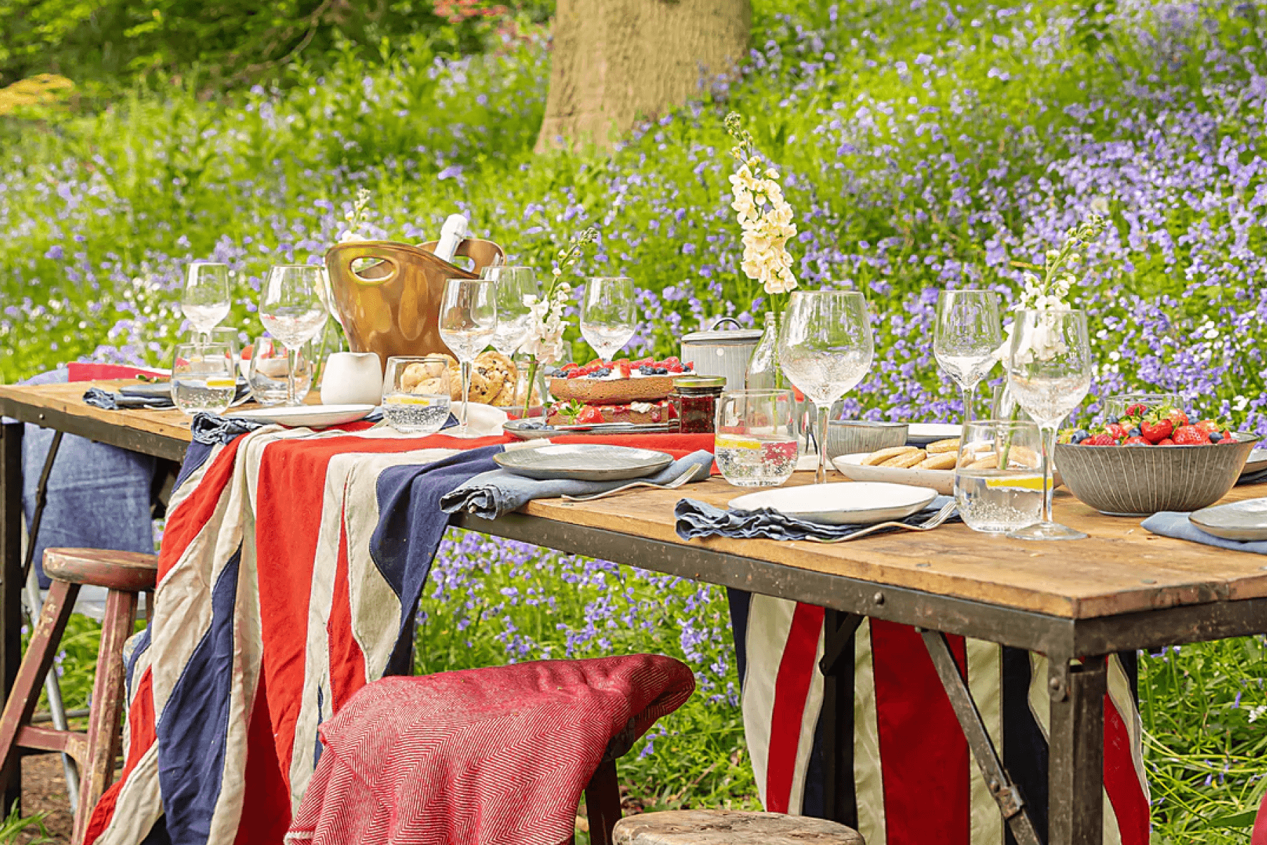 A rustic long table set with wine glasses and dishes is draped with a Union Jack flag in front of greenery and purple wildflowers.