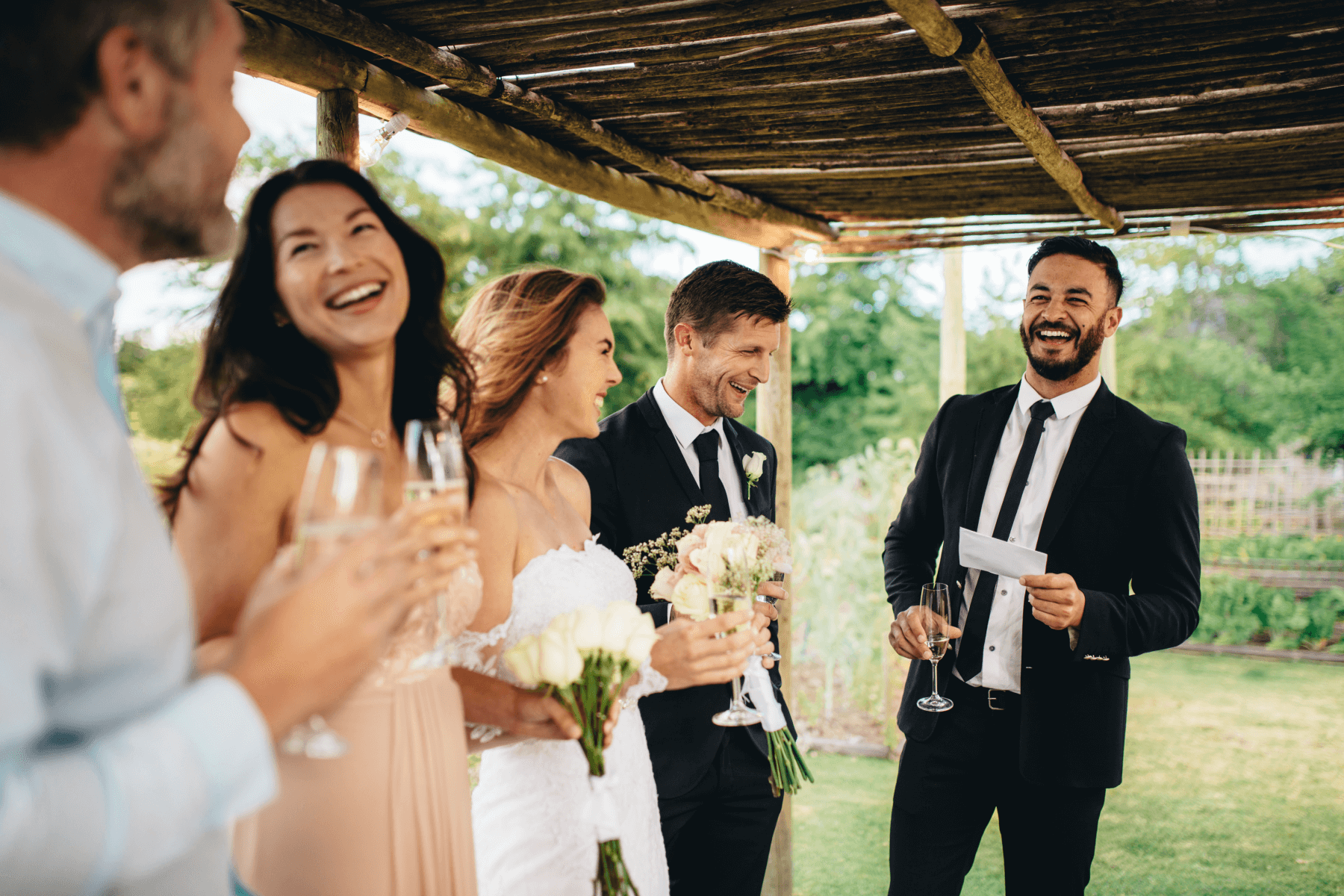 Photo of a well-dressed man giving a speech to a laughing bride and groom and two guests.