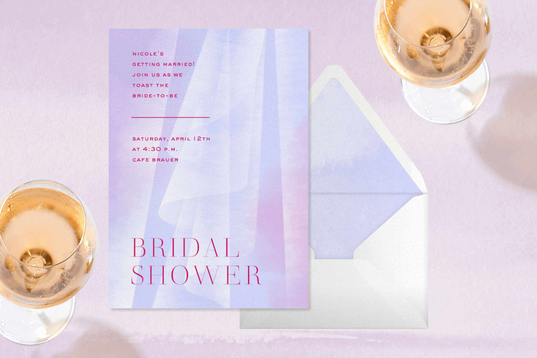 A bridal shower invitation with a light purple, abstract depiction of a bridal veil and white envelope lies beside two rock candy pops and a glass of rosé.