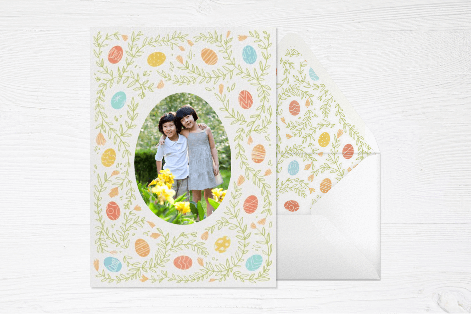 An Easter card with an egg-shaped photo of two young children surrounded by illustrated Easter eggs and green vines, next to a matching envelope on a white wood backdrop.