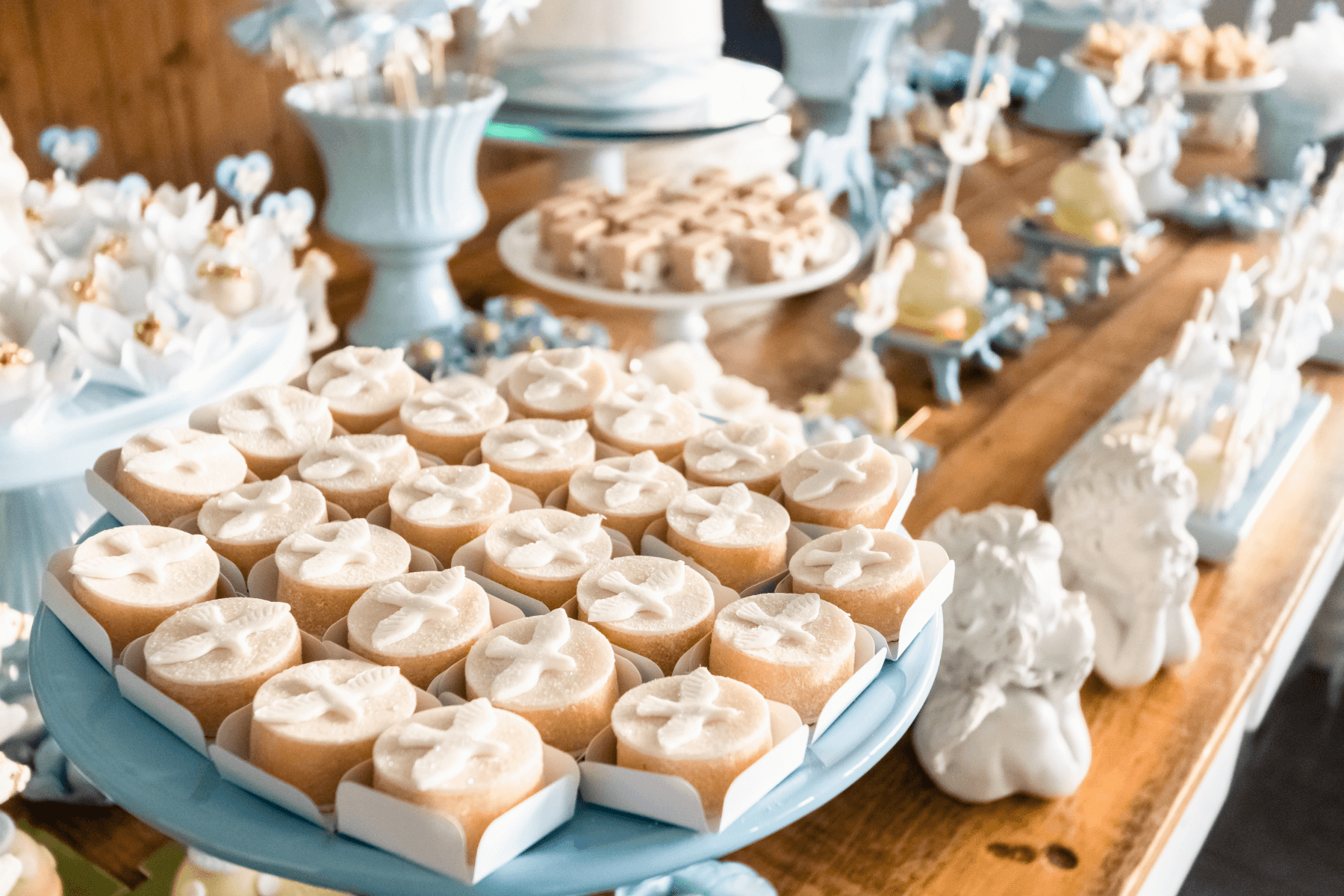 A table topped with blue and white cake stands with pastries featuring a white dove on top, ceramic angels, and blue and white dishes filled with sweet treats.