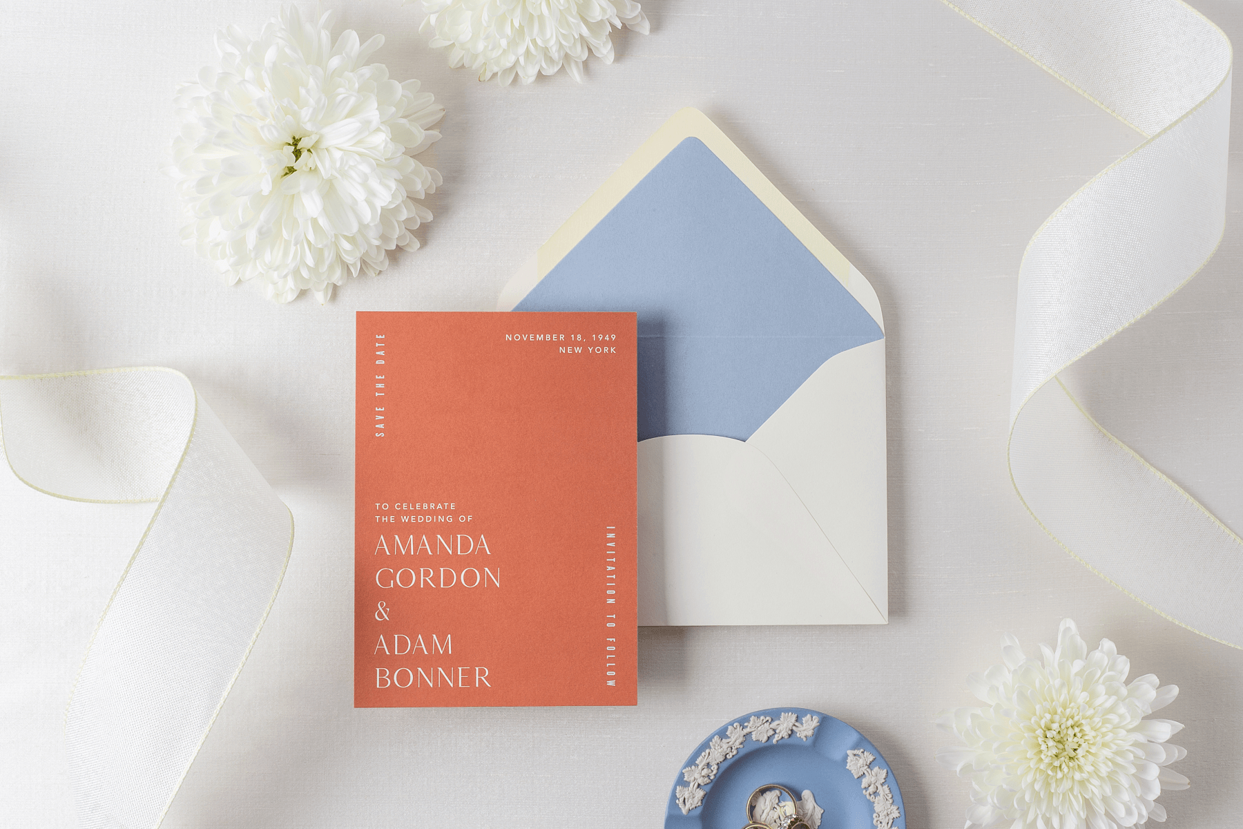 Printed rust-colored save the date invitation sits on top of an open white envelope with a light blue liner. Next to card are white flowers, strips of white ribbon, and a light blue Wedgwood dish holding two wedding rings. 