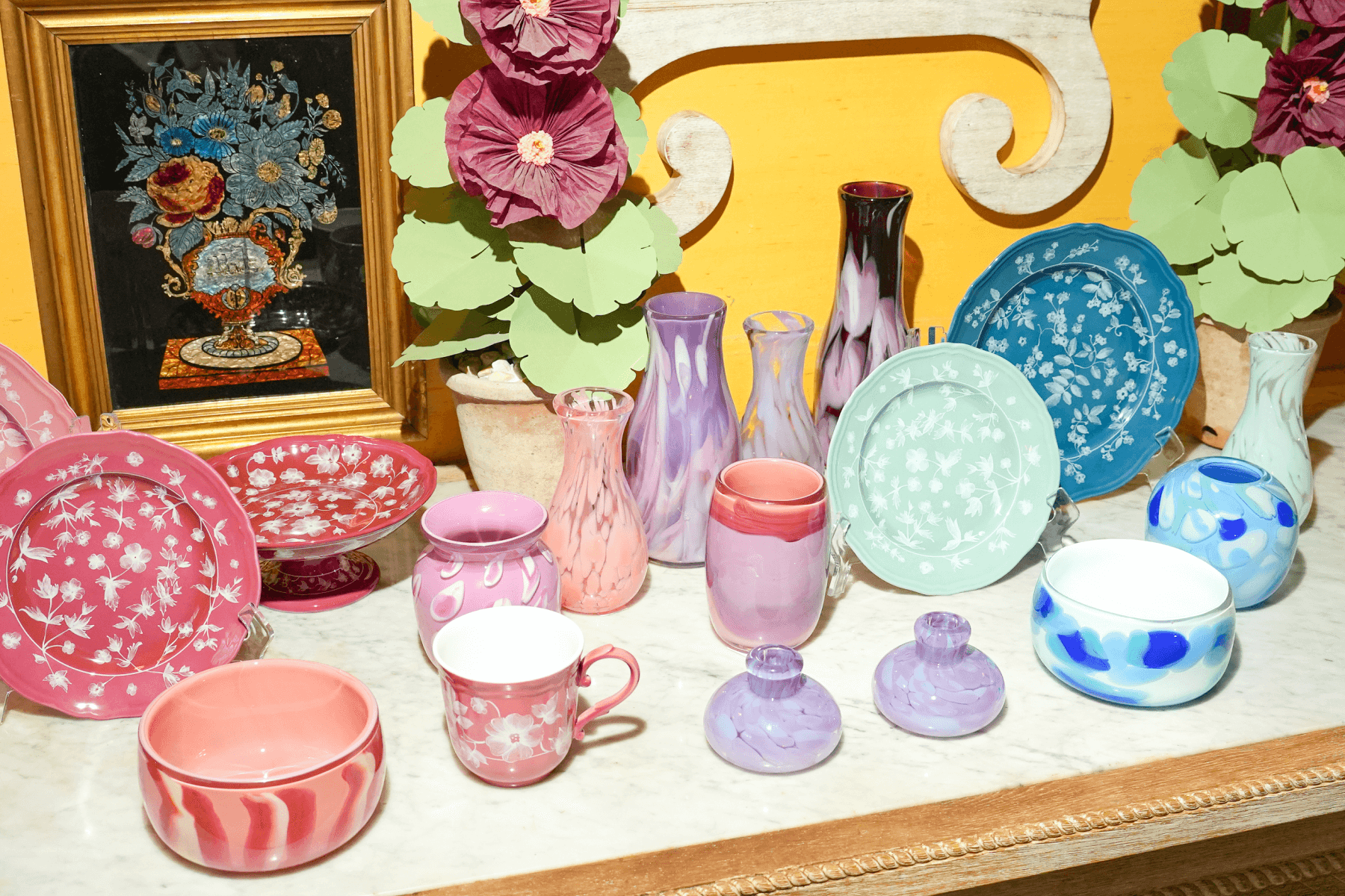 A table topped with colorful tableware, including plates, mugs, and vases in pink, purple, and turquoise, with floral and painted designs. 
