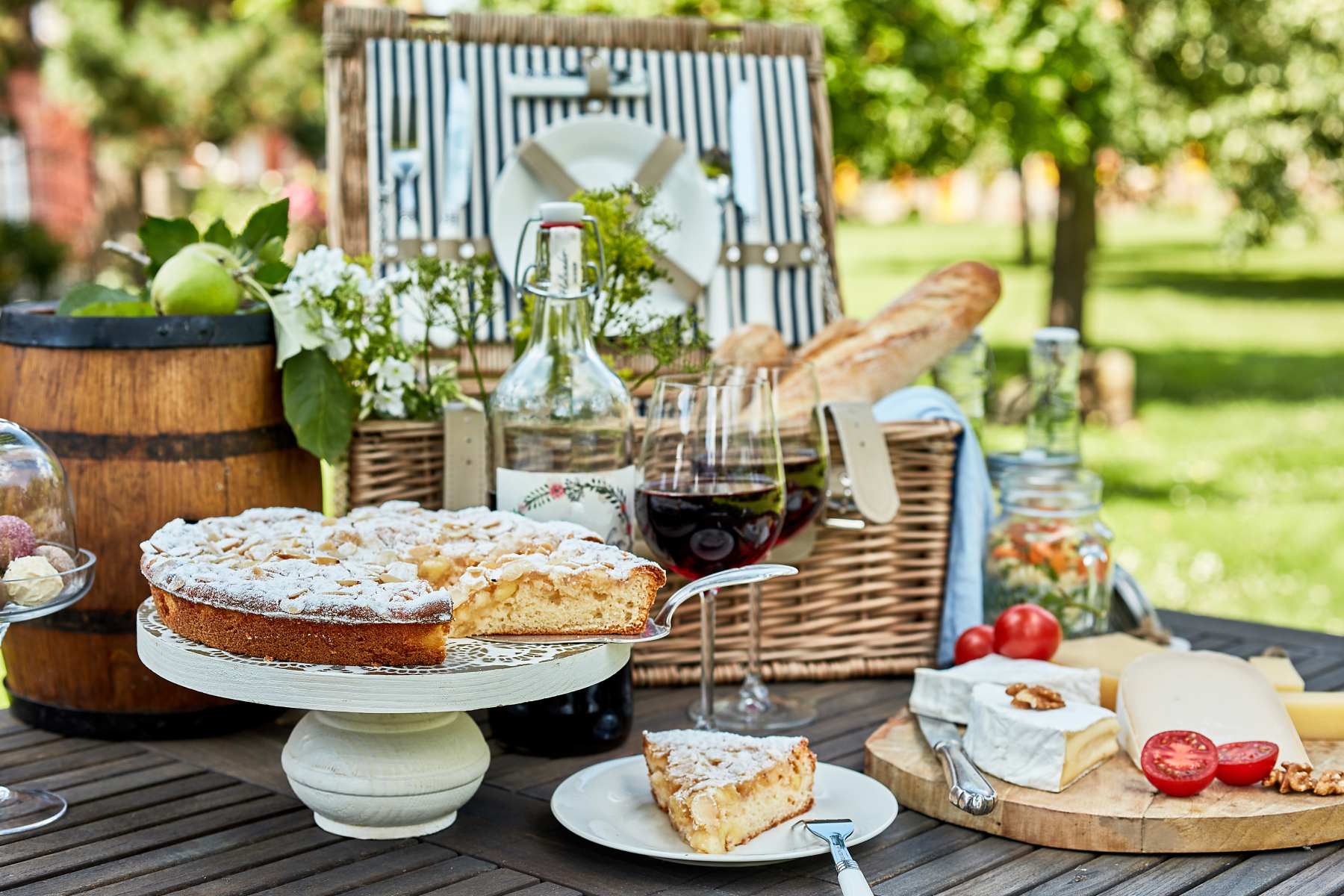 ALT: A wooden outdoor table topped with a picnic basket, baguettes, wine glasses filled with red wine, a circular tart on a serving stand, and a platter of cheese. 