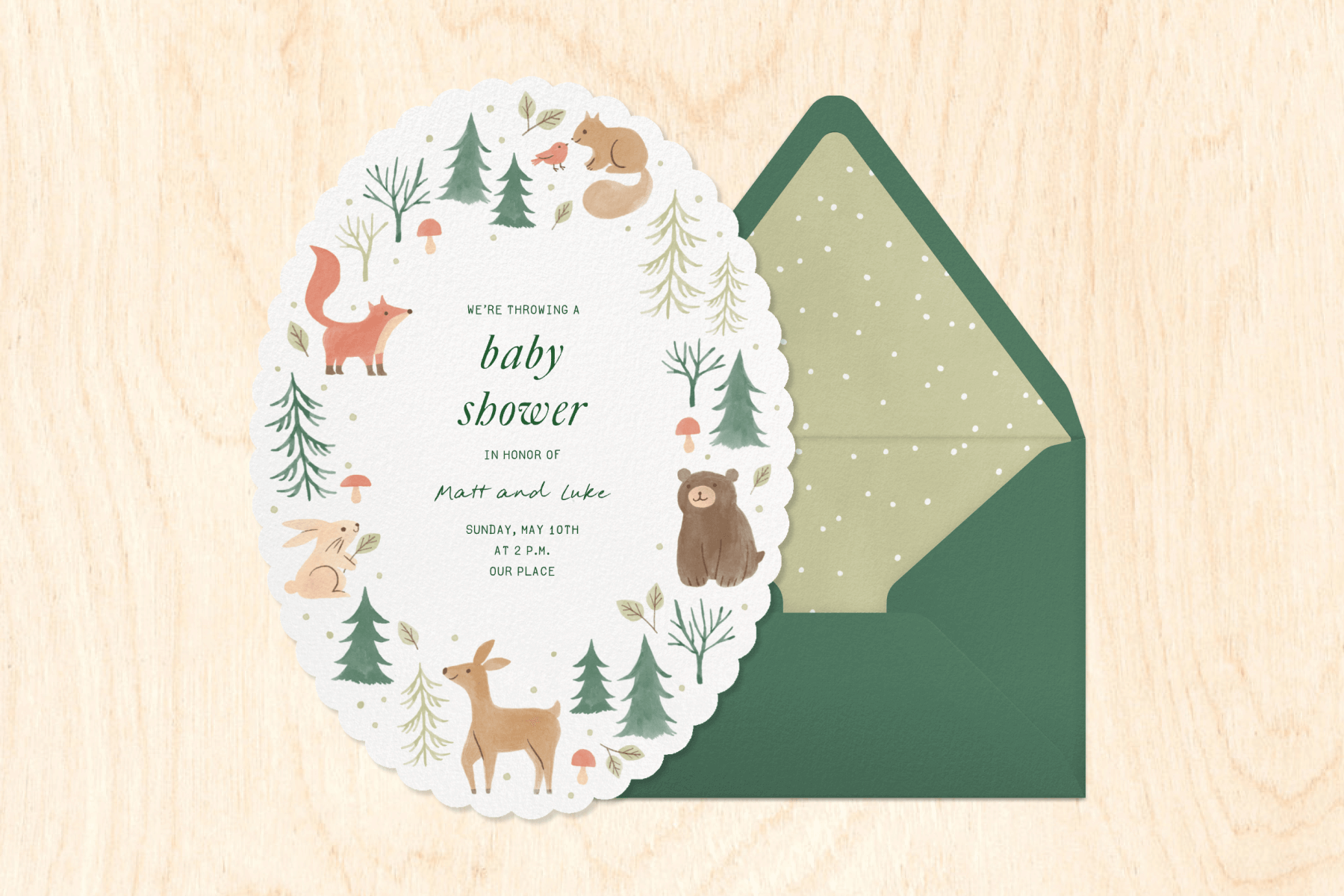 An oval-shaped baby shower invitation with scalloped edges and illustrations of trees and woodland animals, including a deer, a bear, a fox, a squirrel, and a rabbit, next to a green envelope with a light green liner with white polka dots, over a light wood grain backdrop. 