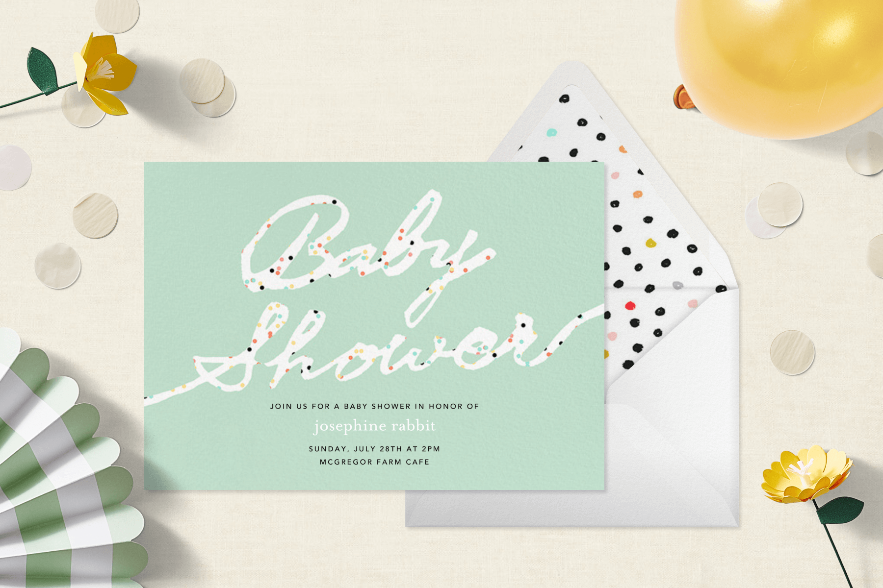 A mint green baby shower invitation with the words “Baby Shower” in white script dotted in rainbow specks, surrounded by yellow paper flowers, a green striped fan, confetti, and a yellow balloon.