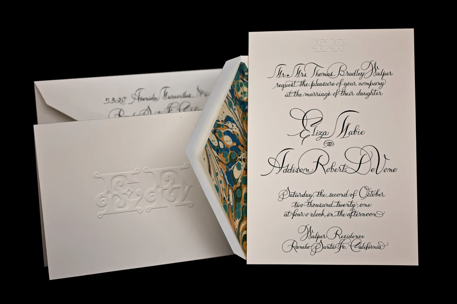 A wedding invitation with heavily embellished black hand-written calligraphy beside an embossed white folded card and an envelope with colorful marbled liner.