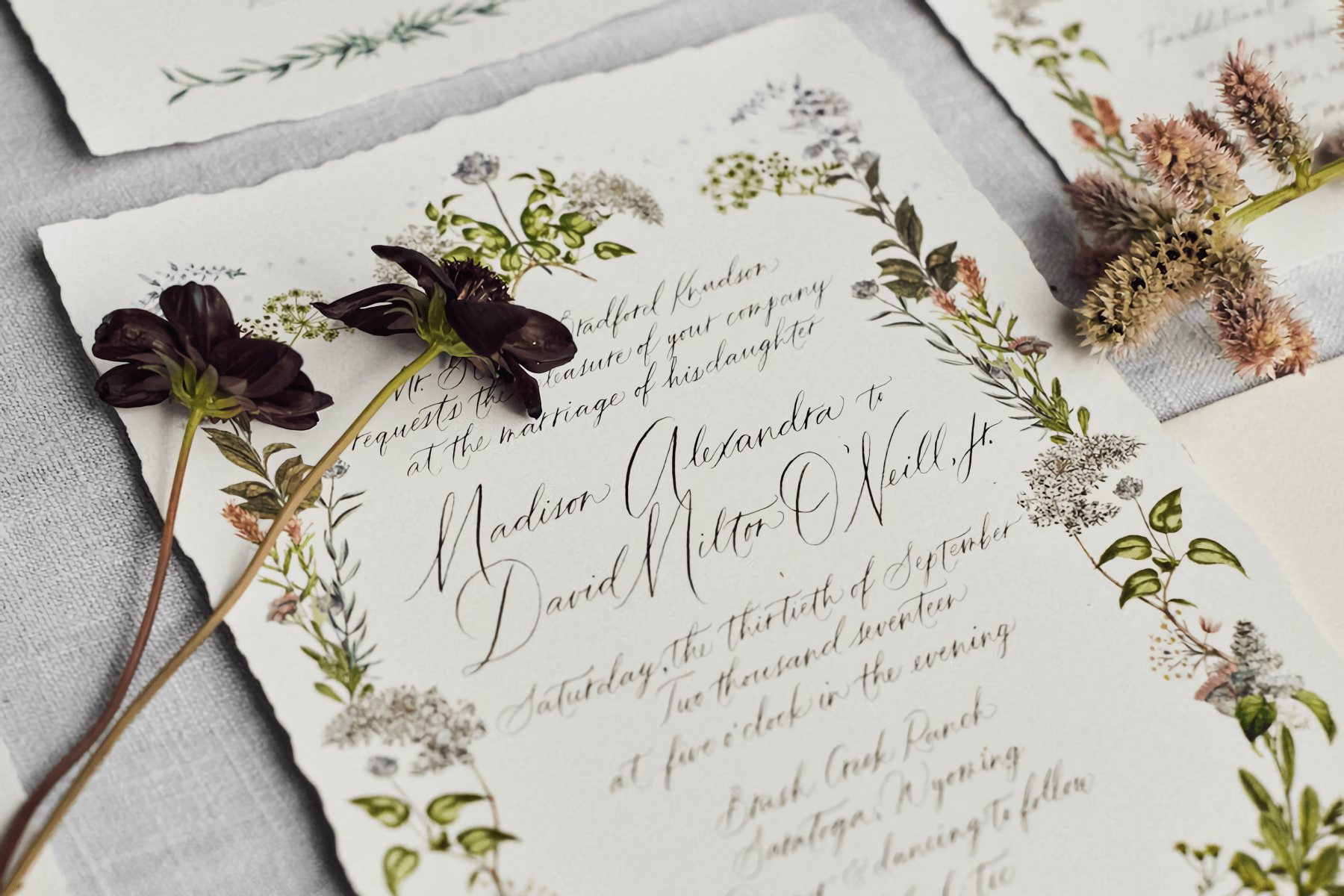 A wedding invitation with organic edges, a delicate floral border, and hand-written calligraphy, with small flowers laid on top.