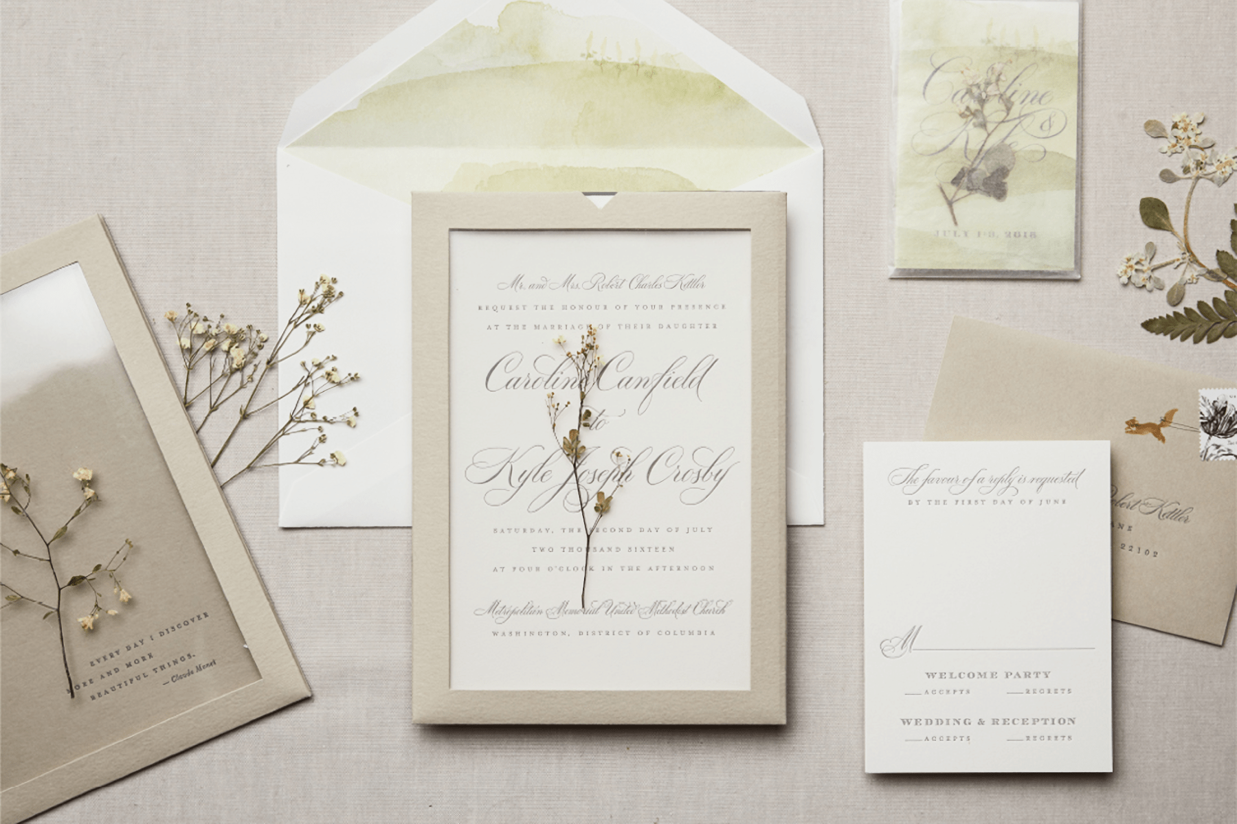 Various elements of a wedding invitation suite with a cream colored text-only invitation, a clear overlay, RSVP card, and envelope and little dried flower stems that will presumably be inserted into the envelopes for mailing.