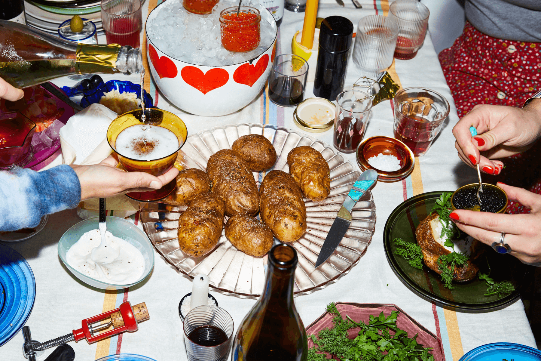 A crowded table top with baked potatoes, caviar on ice, parsley, sour cream, and many mismatched glasses and taper candles, plus the hands of people partaking in the meal.