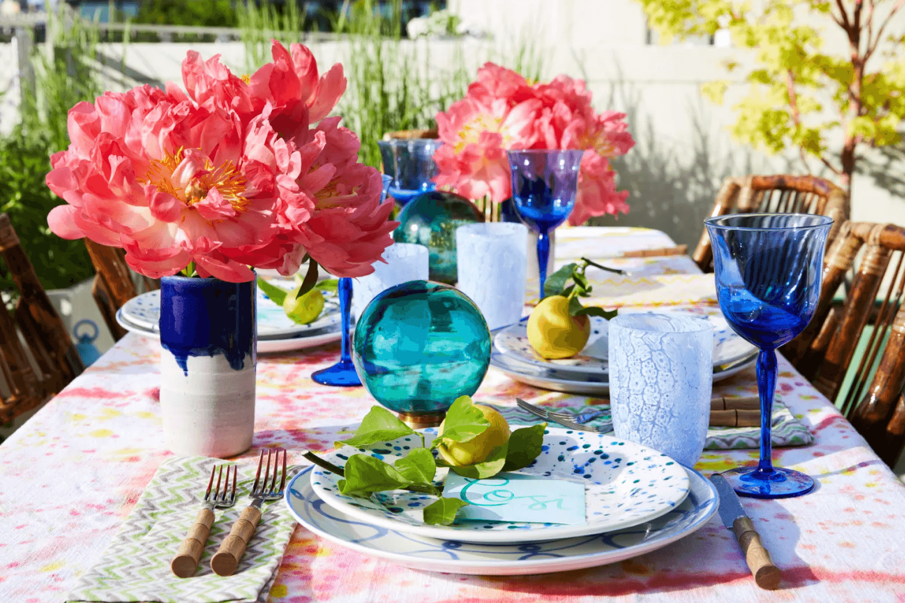 An outdoor table is set with blue glassware, and large pink flowers in vases.