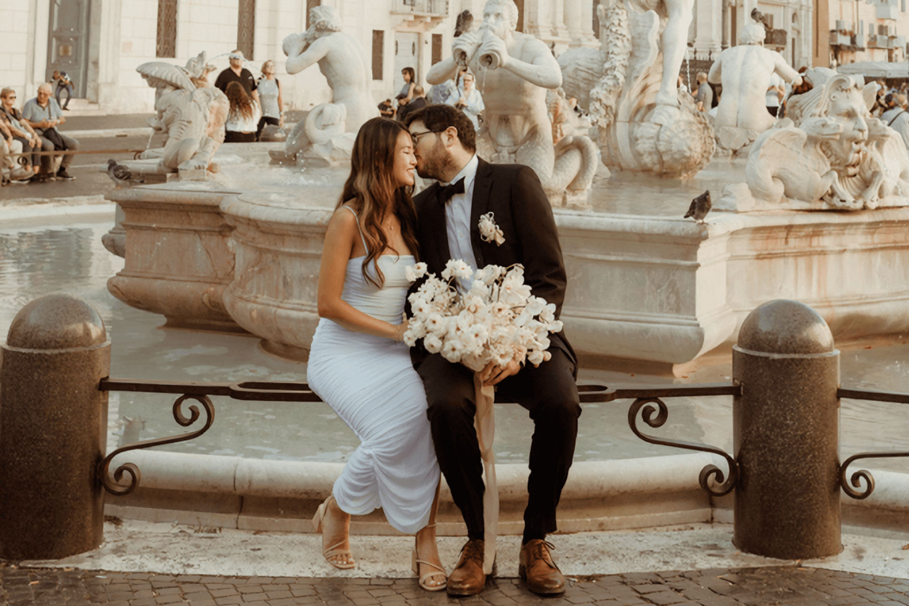 A woman and man in wedding attire and carrying a bouquet of white flowers nuzzle affectionately as they sit on a rail in front of an intricately sculpted fountain.