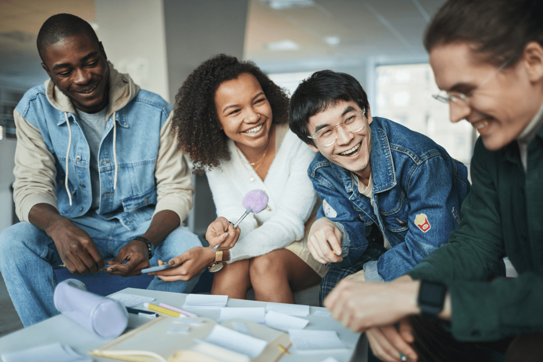 A group of four young people smile as they sit over a table with small note papers, a book, and pens spread out, as if they are collaborating on a project.