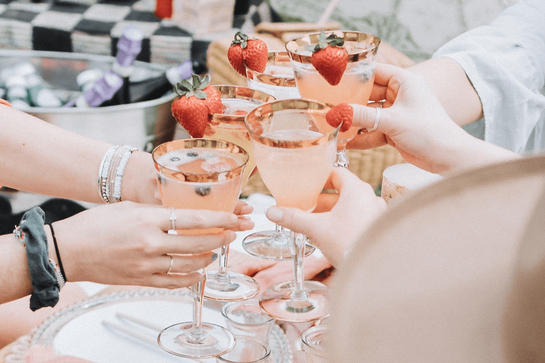 5 hands toasting pink cocktails adorned with berries over a nicely set table.