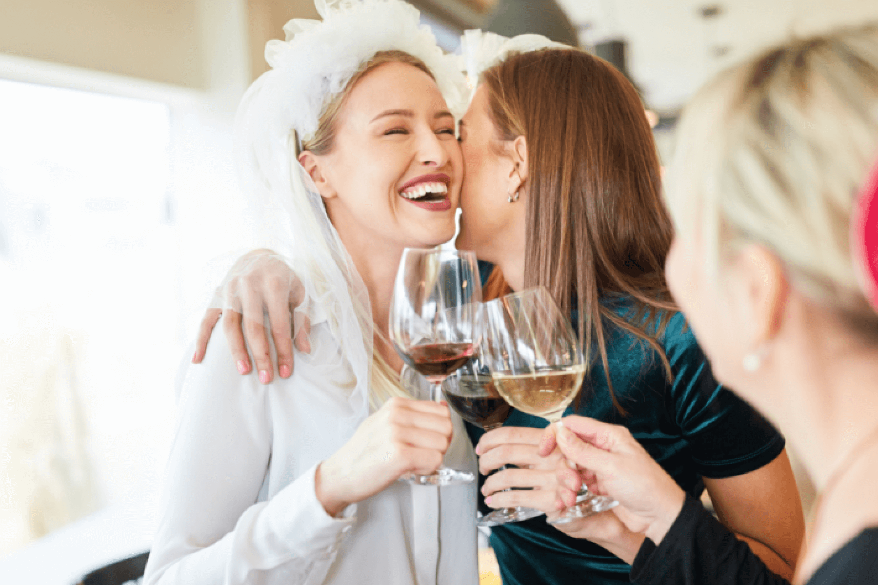 Two women, one wearing a white veil, embrace while clinking wine glasses.