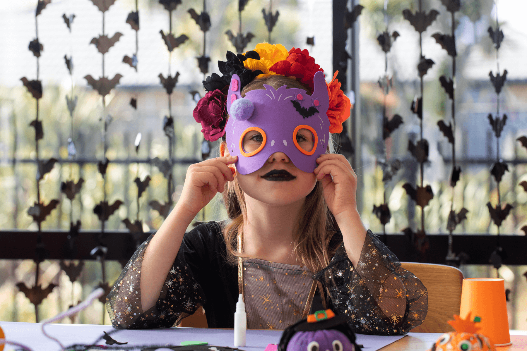 A child in a starry witch costume and a crown of colorful paper flowers holds up a purple foam bat mask at a craft table set with Halloween crafts, in front of a window hung with strands of paper bats.