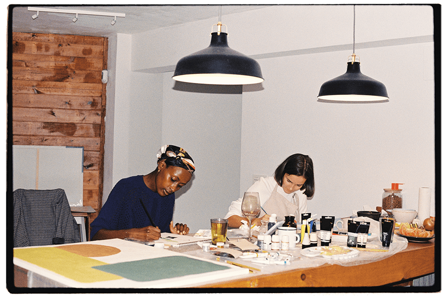 A vintage-looking photograph of two women working on paintings sitting at a high table covered in paint supplies, a canvas, and a wine glass, with two pendant lights hanging above them. 