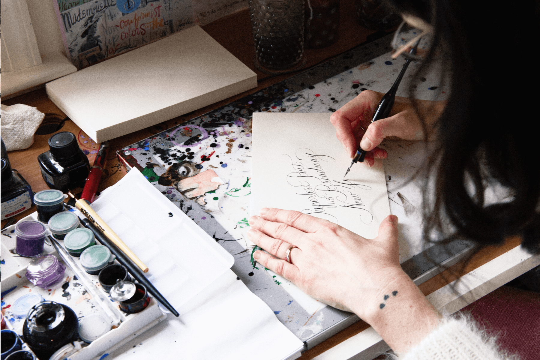 Stephanie Fishwick sitting at her desk writing calligraphy, surrounded by paint and ink.