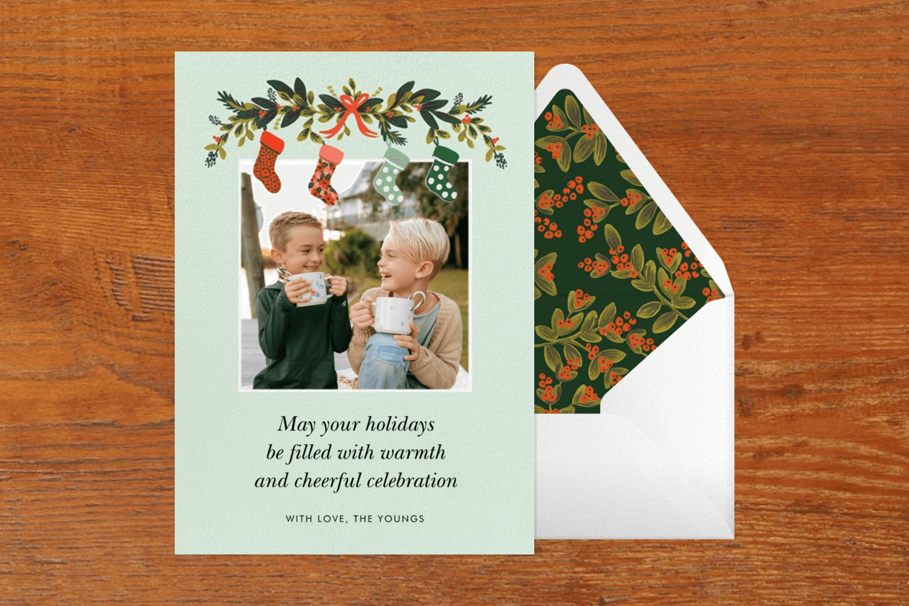 A mint-colored photo holiday card with an illustration of stockings on a garland, featuring a photo of two boys drinking hot cocoa.