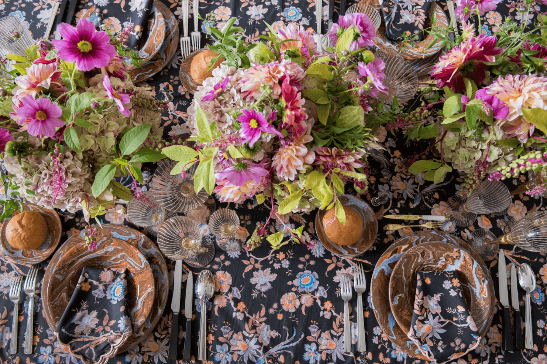 Wide crop of a dinner table set with a black floral tablecloth, maroon marbled plates, and purple flowers. 