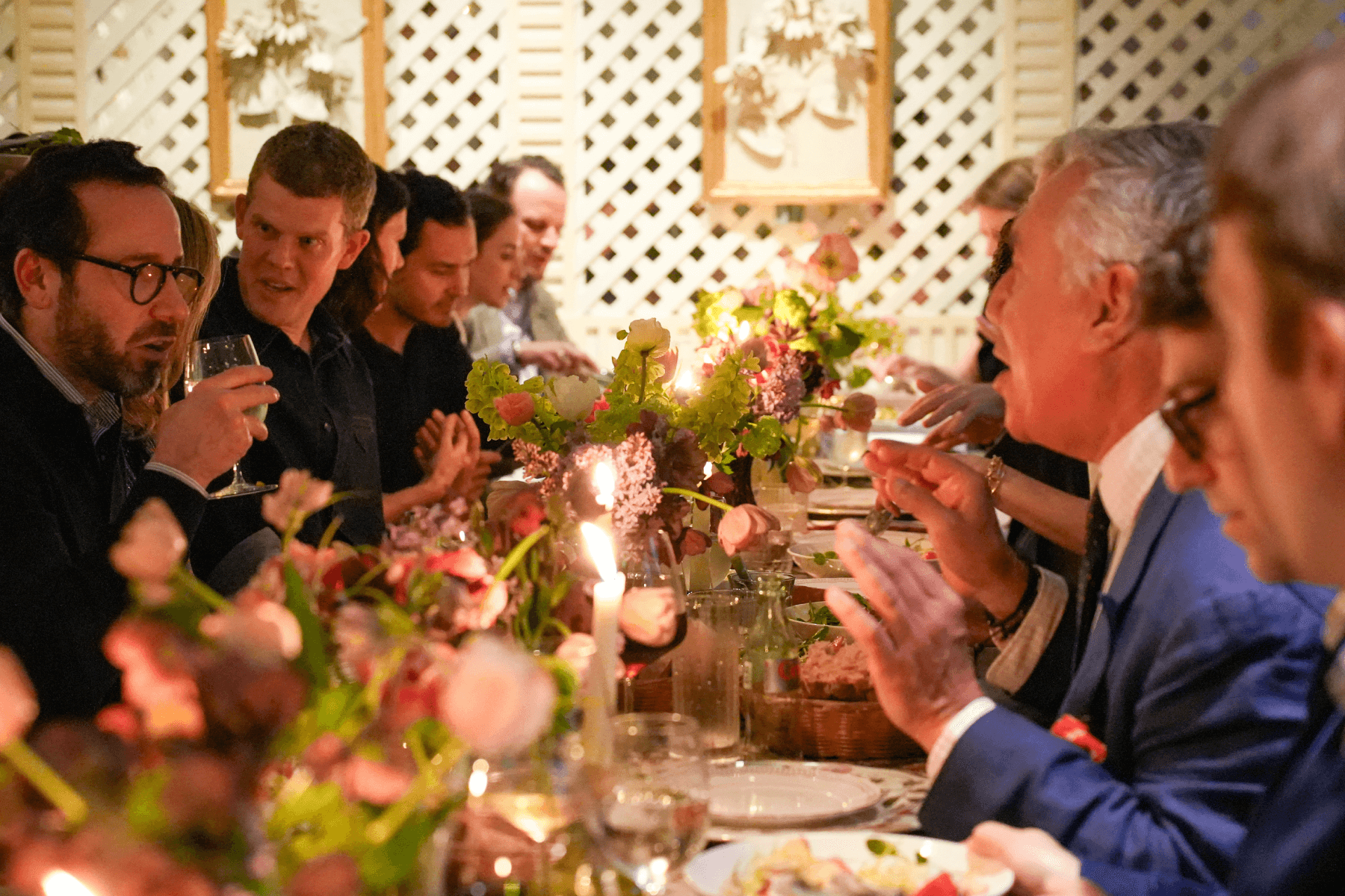 A dining table topped with candles and florals, with guests drinking, dining, and chatting. 