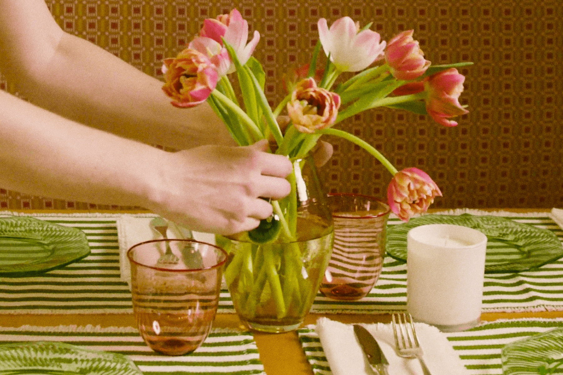 ALT: Hands are arranging pink flowers in a vase in the middle of a dining table set with green and white striped place mats, green plates, pink glasses, and a candle.