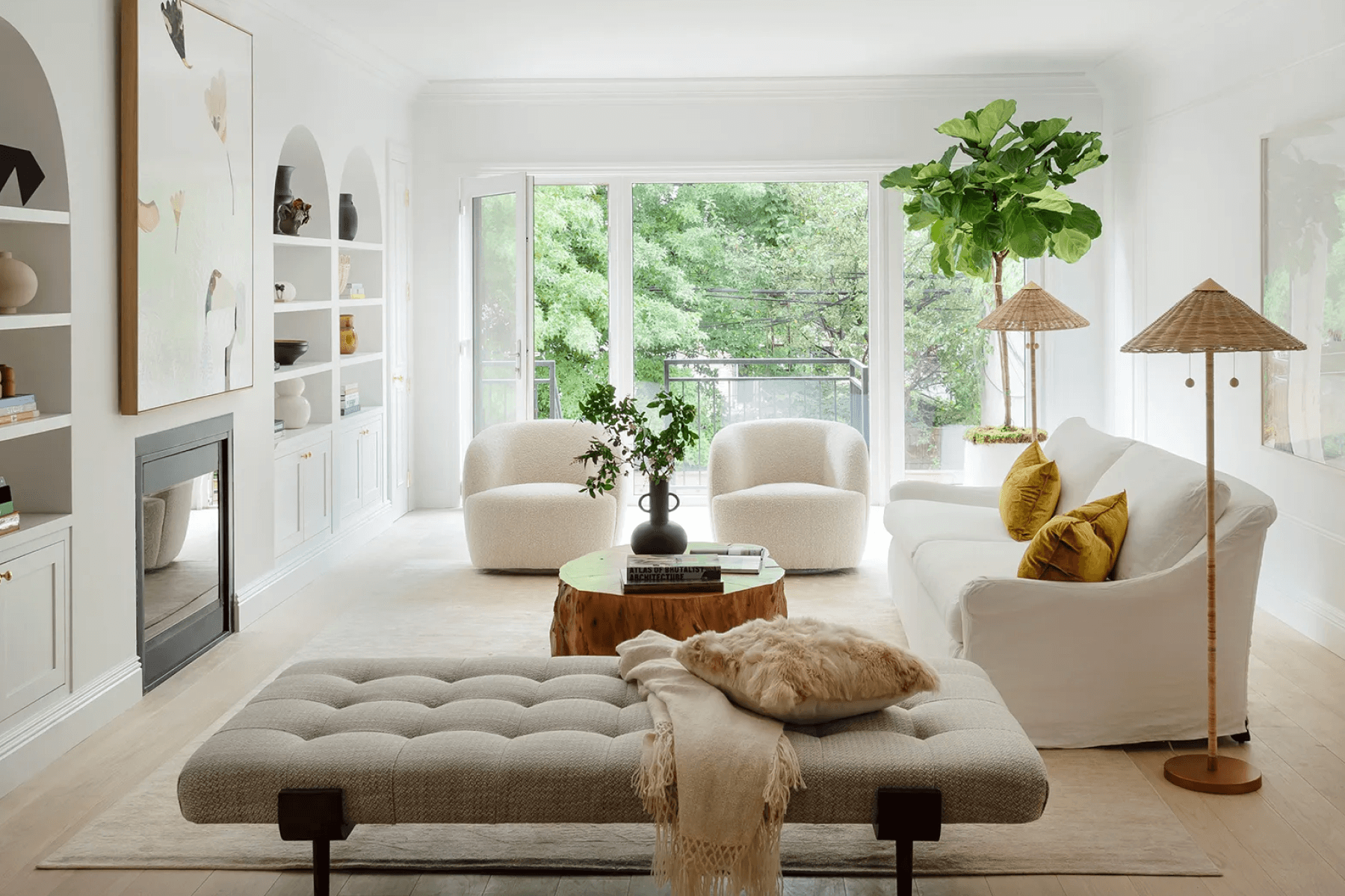 An airy living room with a tufted chaise lounge, two white modern arm chairs, a white couch, two rattan floor lamps, a fiddle leaf fig, and a fireplace with built-in arched shelves.